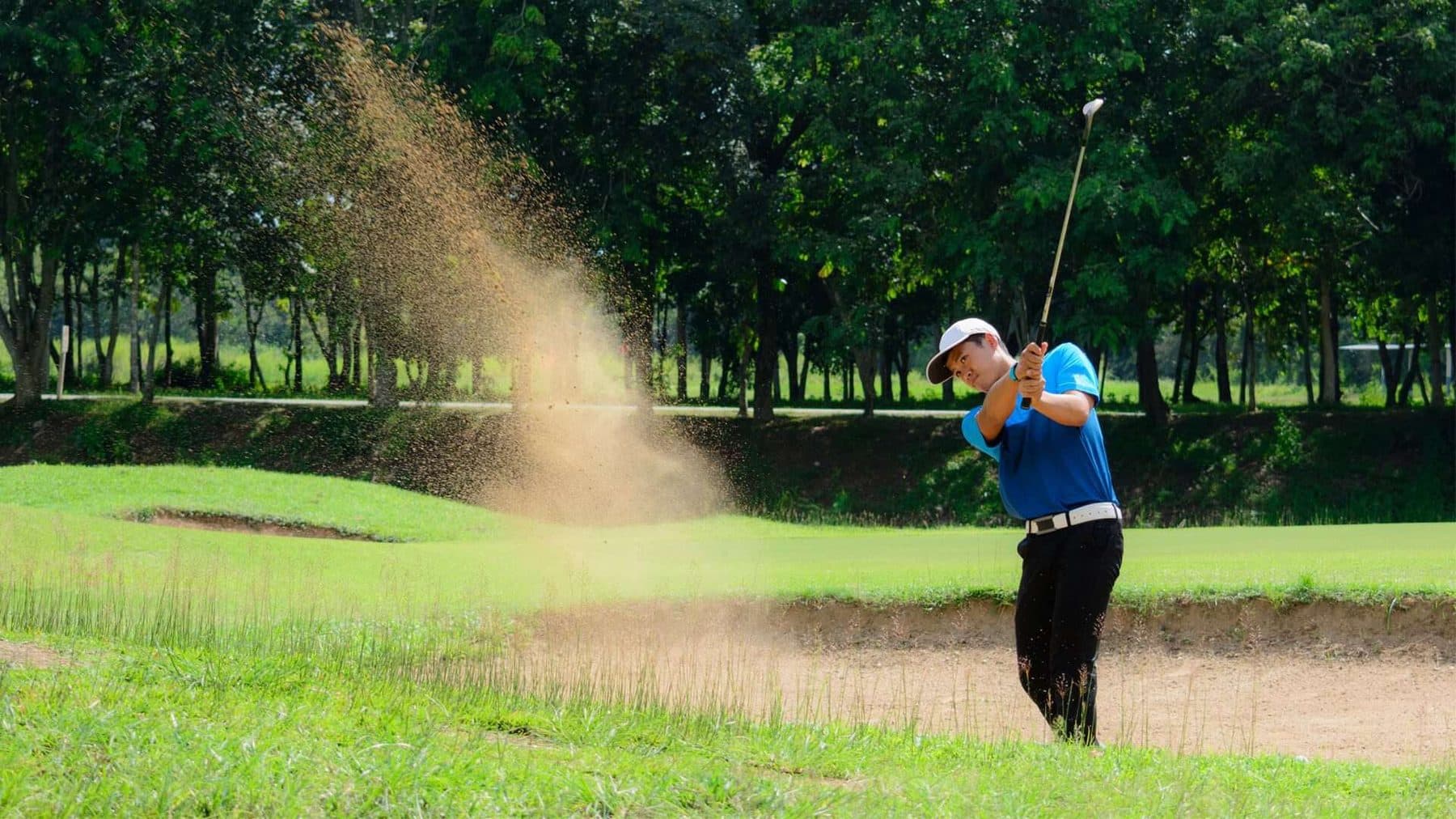 A golfer splashing out of a greenside bunker, trees lining the fairway behind