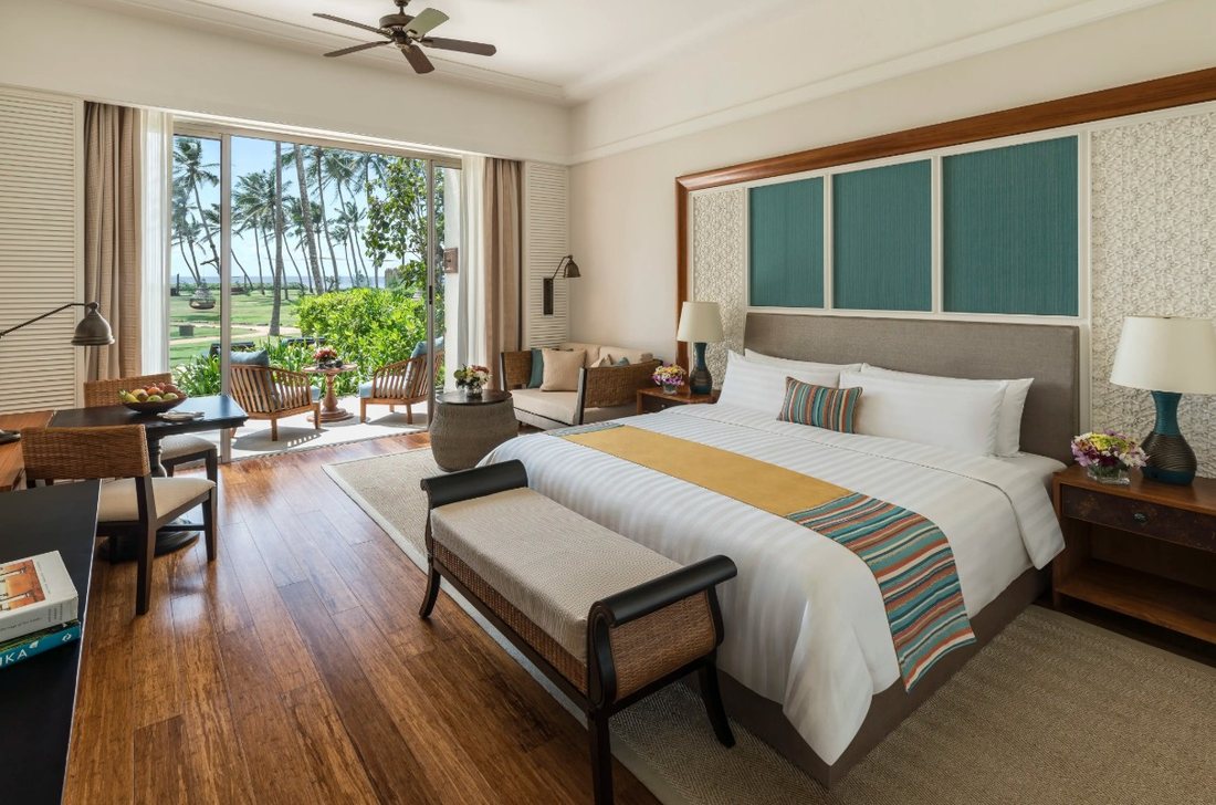 A deluxe hotel bedroom with a large bed, a ceiling fan and doors open onto a balcony with palm trees