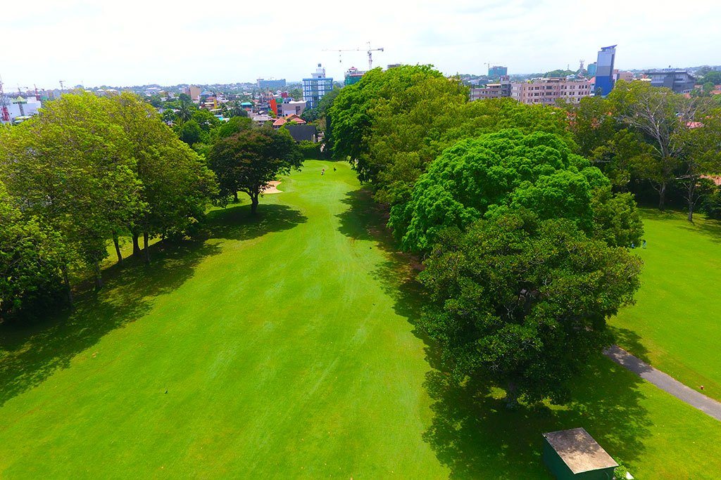 A tree-lined fairway at Royal Colombo Golf Club seen from above, with the Colombo skyline beyond