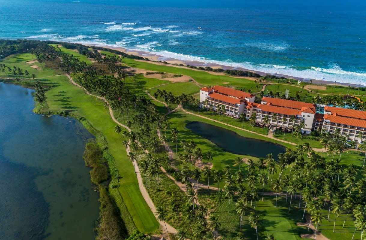 Aerial view of the Shangri-La Hambantota golf course running along the Indian Ocean, with the resort's red-roofed buildings, a lagoon and palm trees